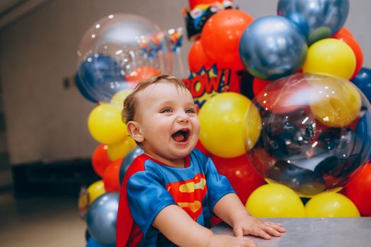 Smiling boy in superhero costume surrounded by vibrant balloons indoors.