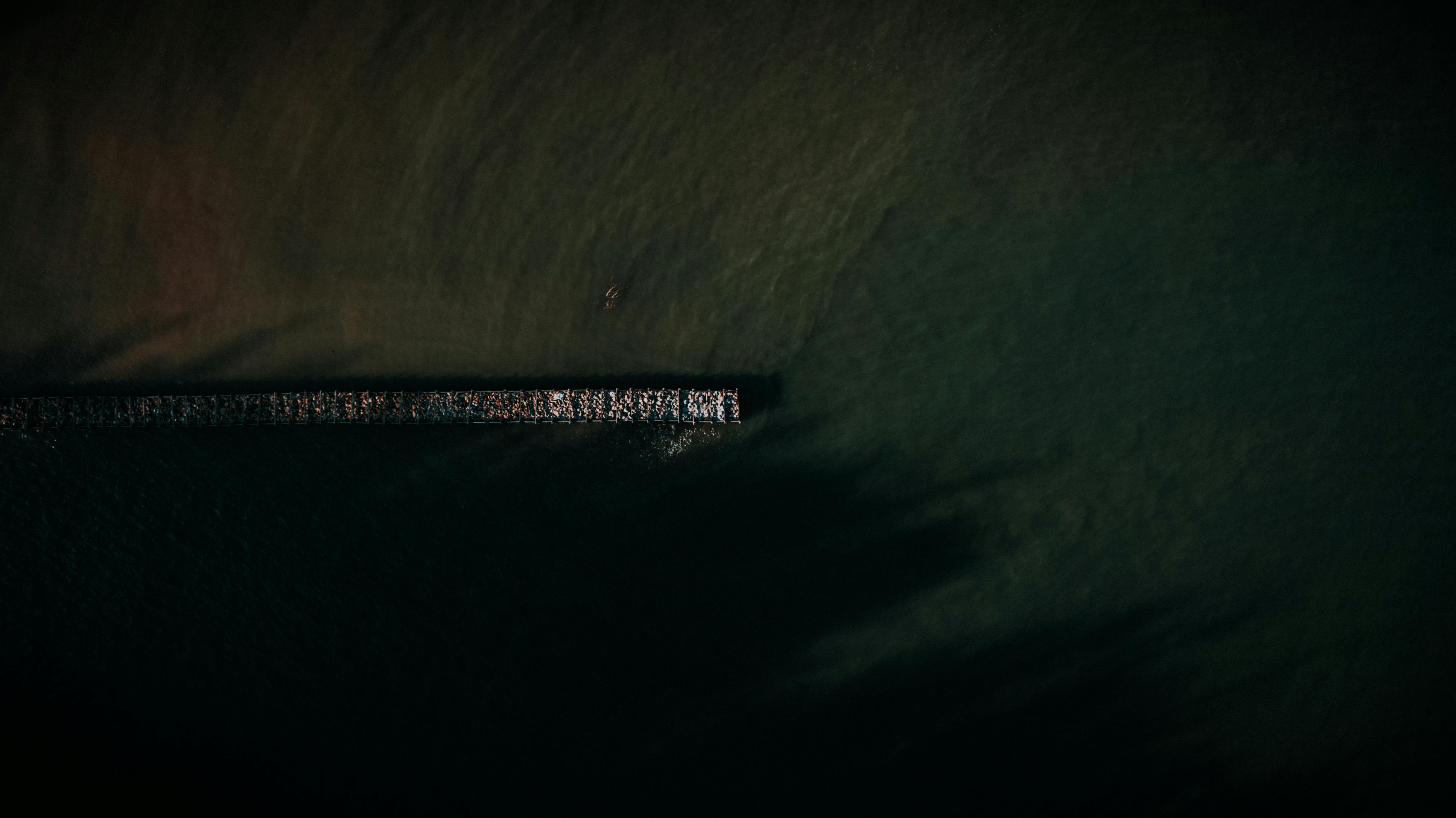 A dramatic aerial view showcasing a pier extending into the dark sea with a lone kayak nearby.