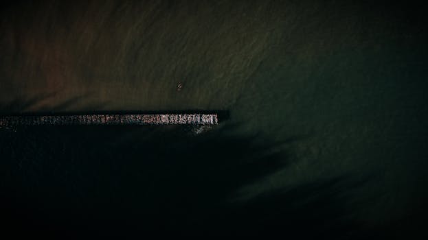 A dramatic aerial view showcasing a pier extending into the dark sea with a lone kayak nearby.