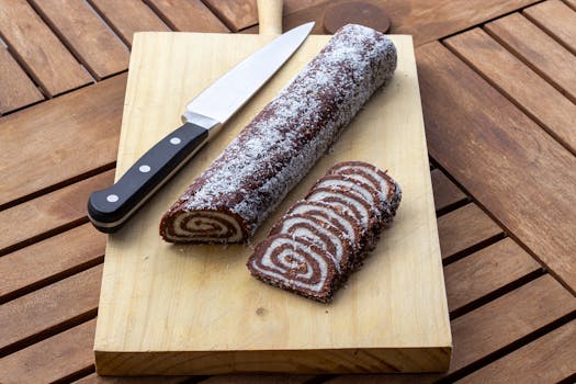 Tempting chocolate Swiss roll with coconut topping, sliced on a wooden board with a knife.
