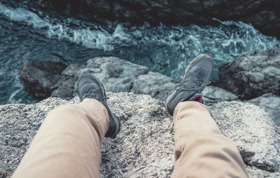 A person sits on a cliff edge, overlooking the ocean with visible feet and shoes, enjoying nature.
