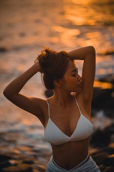 Woman in white bikini posing by the ocean at sunset with serene expression.