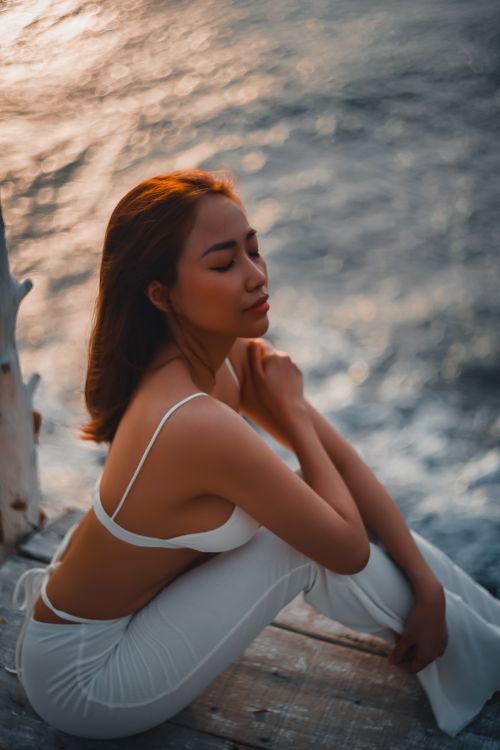 Free A woman in a bikini top and white pants enjoys a peaceful moment by the sea at sunset. Stock Photo
