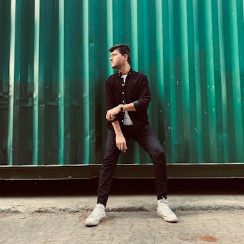 Stylish man in a black outfit poses confidently against a green wall outdoors.