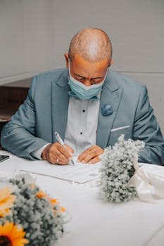 Man in a blue suit signs a document at a formal event with flowers.