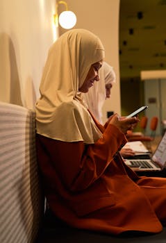 Side view of a woman in a hijab using a smartphone, sitting indoors.