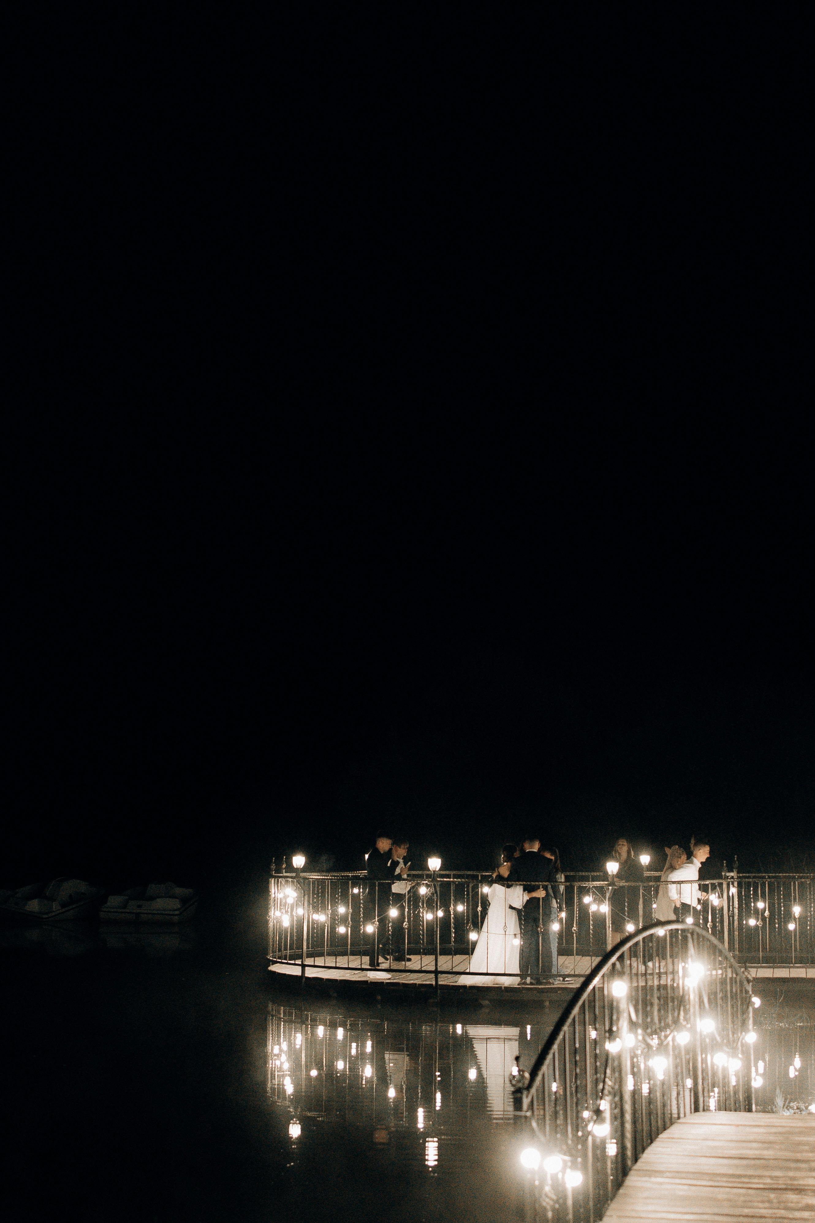 A romantic nighttime wedding with couples on a beautifully illuminated bridge reflecting in water.