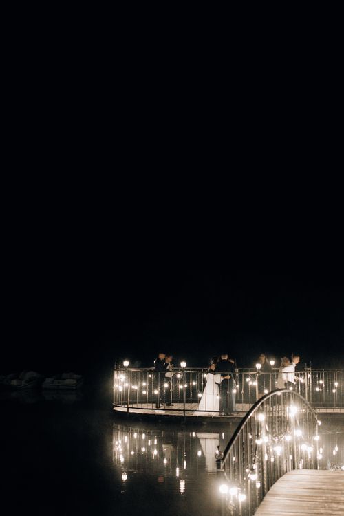 Free A romantic nighttime wedding with couples on a beautifully illuminated bridge reflecting in water. Stock Photo