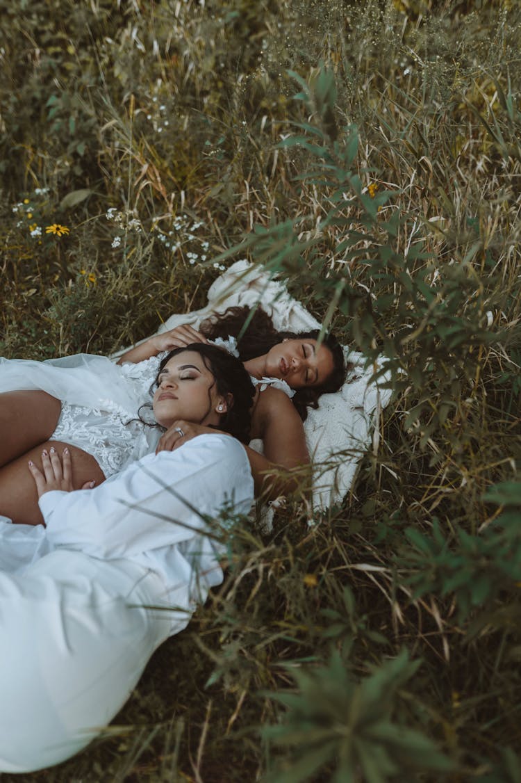 Lesbian Couple In White Dresses Laying On Grass Cuddling