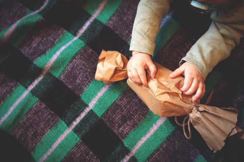 Free A child unwrapping a brown paper gift on a colorful checkered blanket. Stock Photo