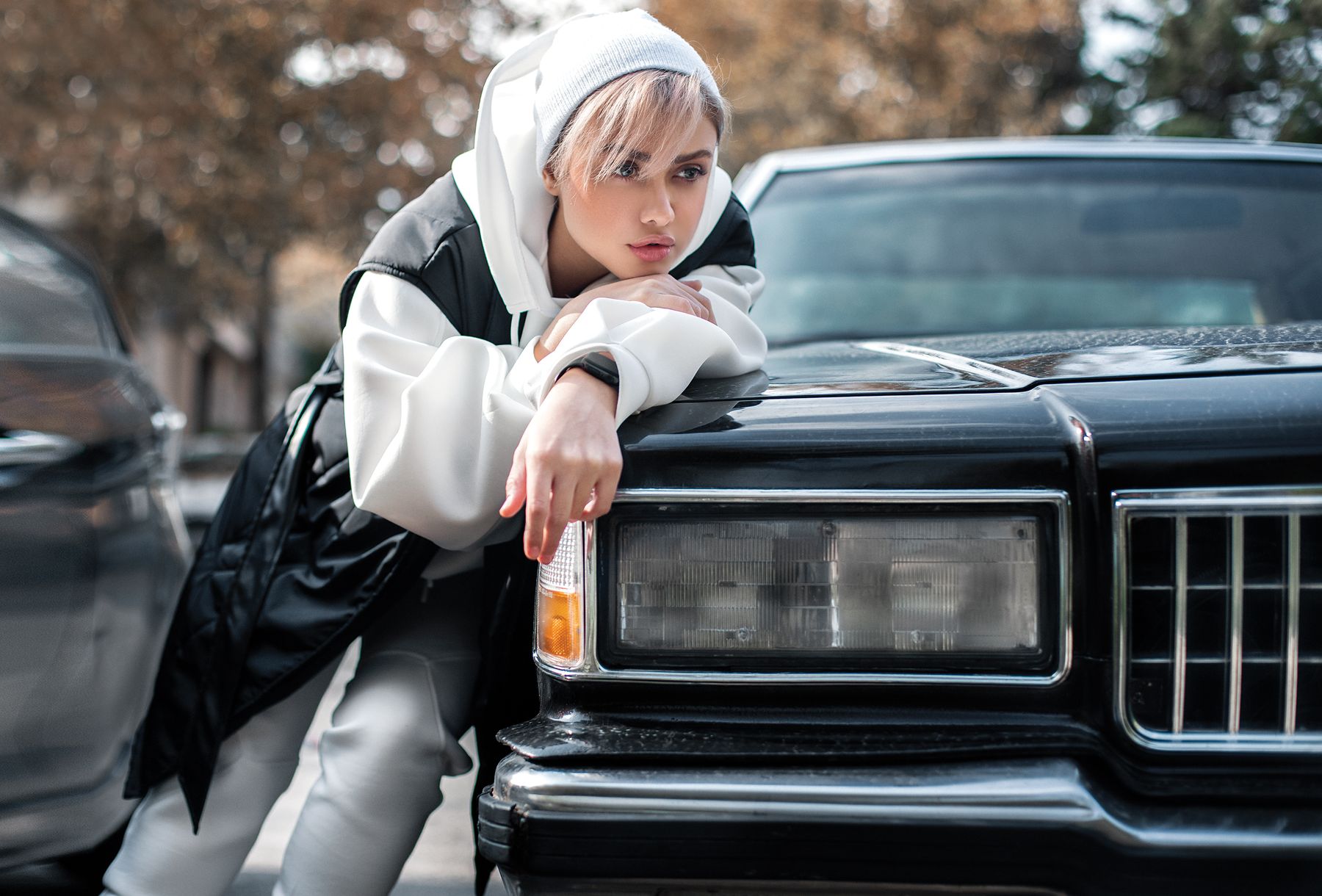 Woman wearing a beanie and oversized hoodie beside a vintage black car