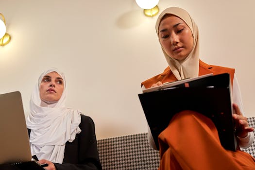 Two women in hijabs collaborating at the office, engaged in work with laptops and notes.