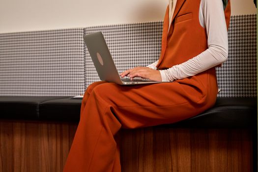 Side view of a woman in an orange suit working on a laptop in a modern office setting.