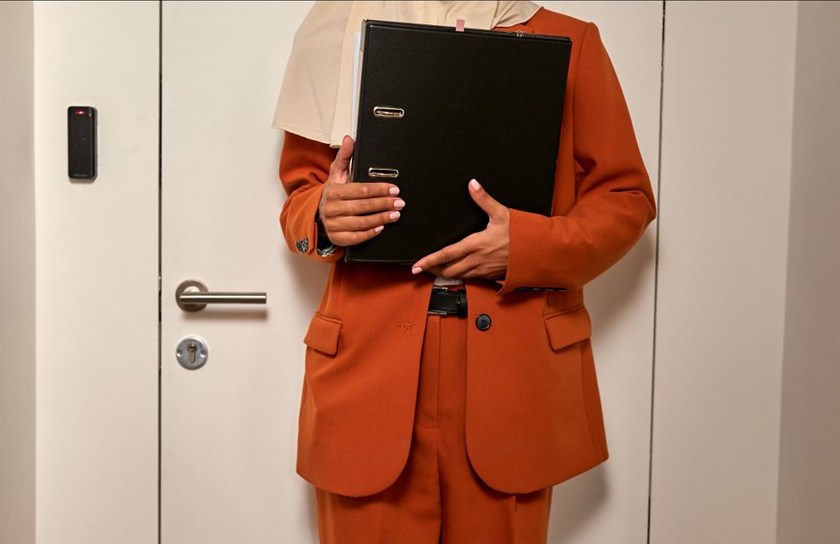 Primer plano de una mujer con traje naranja sosteniendo documentos, en un entorno de oficina.