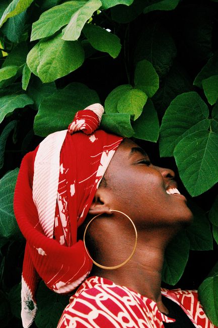 A joyful woman in a red headscarf smiles surrounded by lush green leaves, showcasing natural beauty.