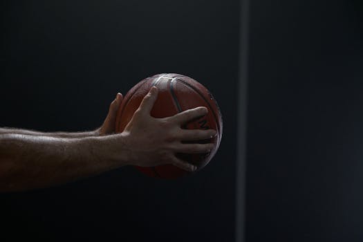 Dramatic close-up of a basketball player's hands holding a ball against a dark background.