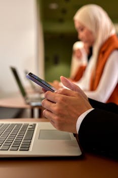 Two women using smartphones and laptops in a modern office setting, emphasizing multitasking and technology.