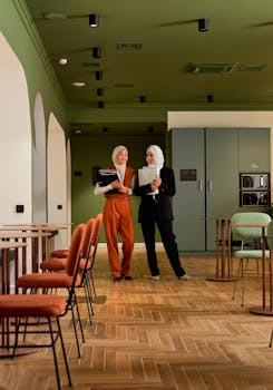 Two businesswomen in hijabs discussing work in a stylish office space with modern decor.