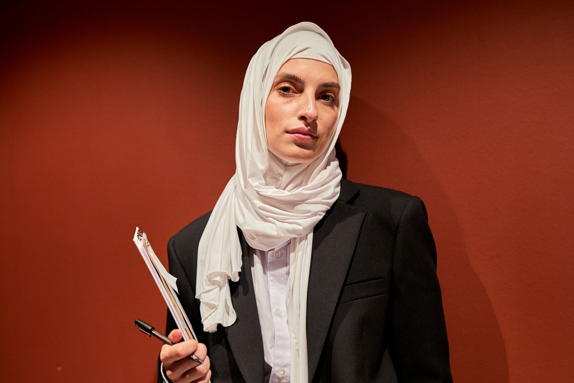 A confident Muslim woman in a hijab stands in a modern office setting.