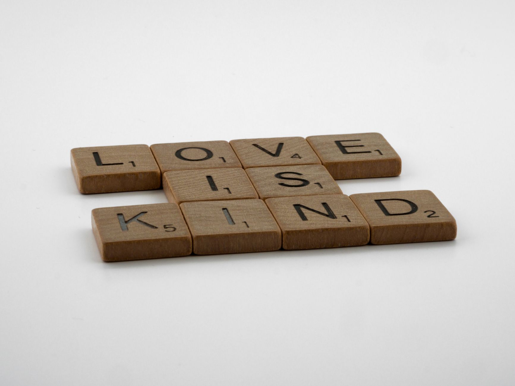 Wooden Scrabble tiles arranged to say 'Love is Kind' on a plain background.