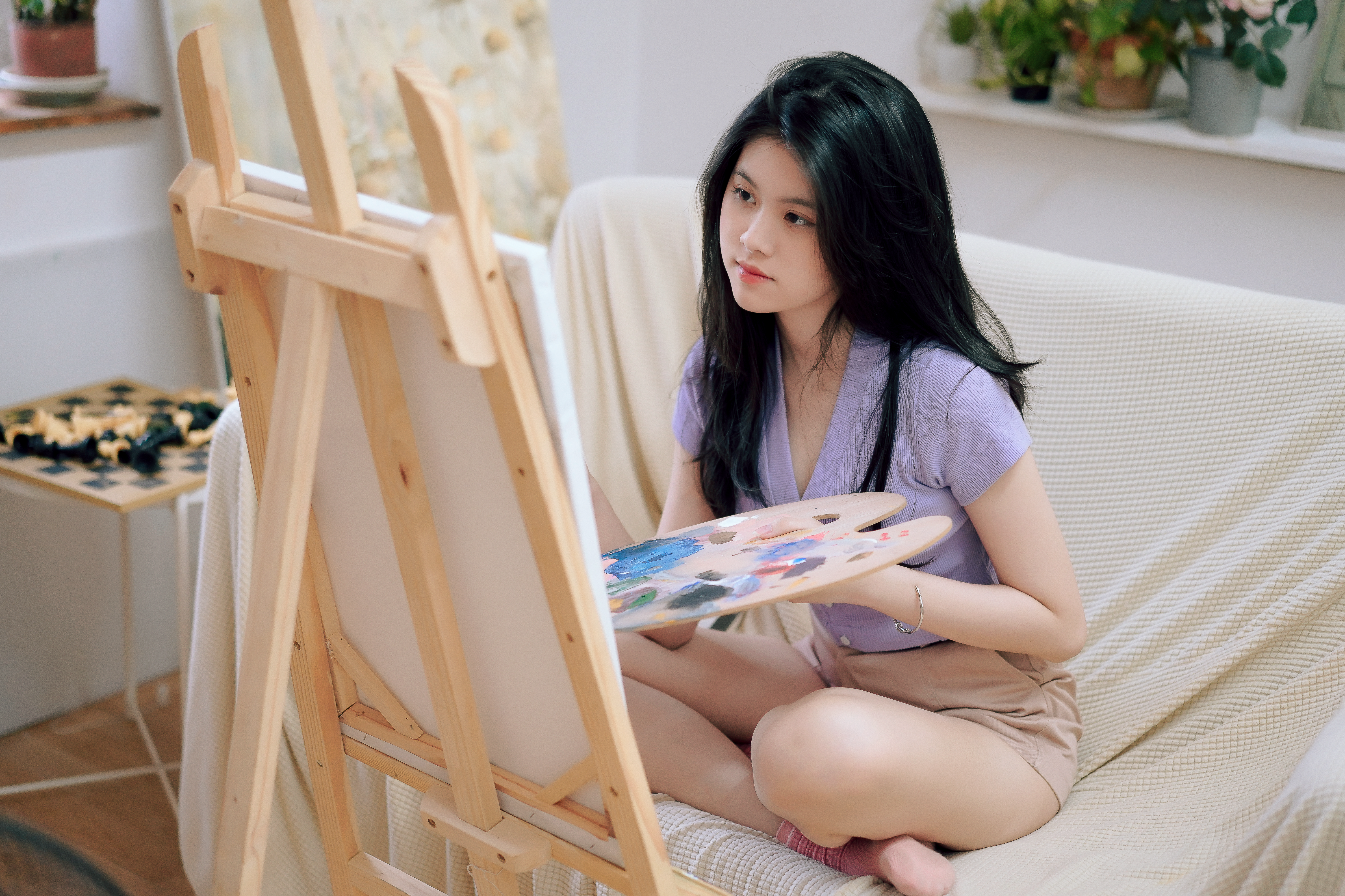 A young woman painting with a palette, sitting in a cozy room with an easel.