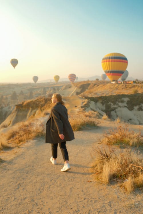A Woman Walking on Dirt Path Near Hot Air Balloons