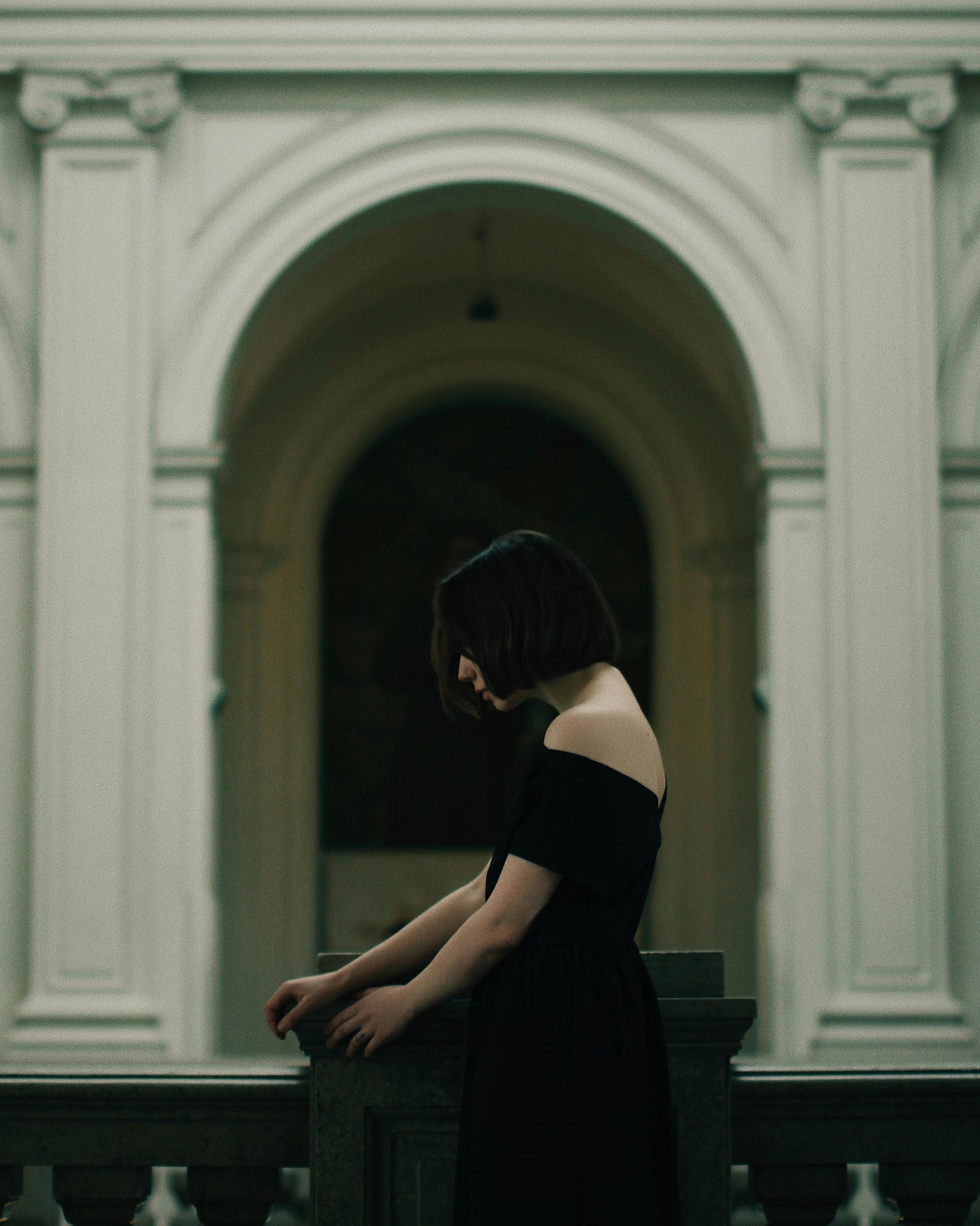 A woman in a black dress stands near classical architectural arches with blurred background.