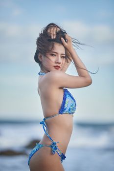 A young woman in a colorful bikini poses by the ocean on a sunny day.