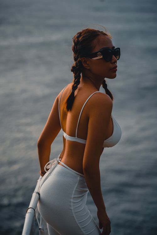 Free A woman in a white outfit and sunglasses enjoys the sunset by the ocean, capturing leisure and style. Stock Photo