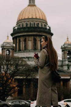 A woman in a brown coat standing with flowers near St. Isaac's Cathedral, showcasing autumn vibes.