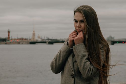 Free Thoughtful woman in a coat by the riverside with a cityscape in the background. Stock Photo
