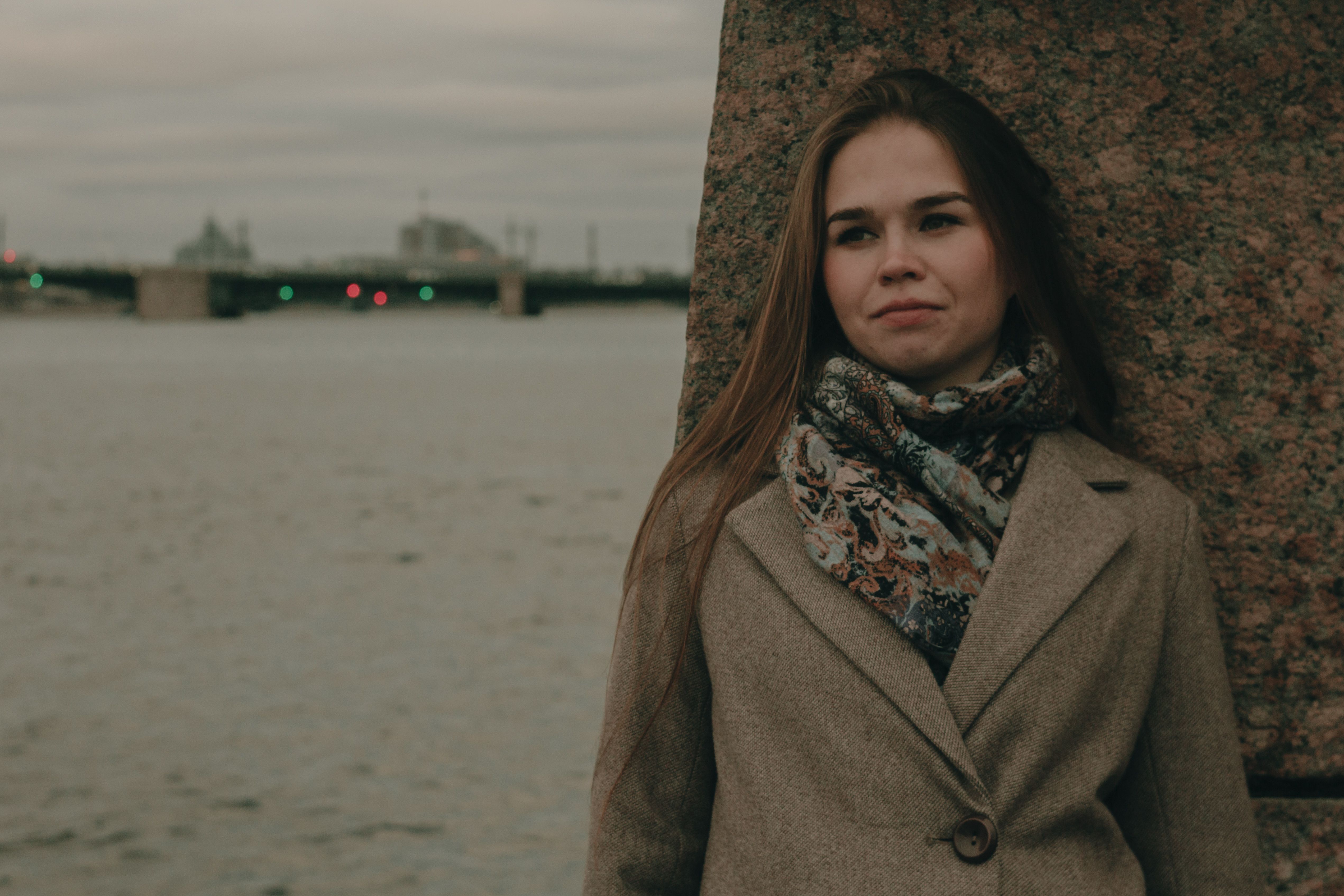 A woman in a brown coat leans against a wall by the river on a cold winter day.