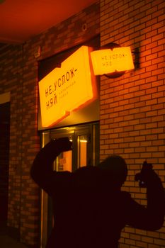 A person poses in front of illuminated Russian neon signage against a brick wall at night.