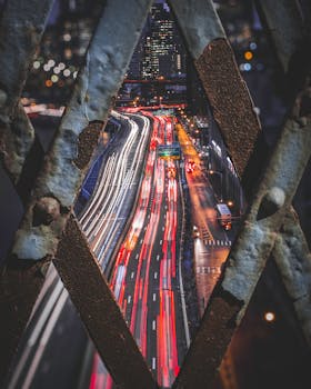 Dynamic night photo of city traffic creating vibrant light trails through a lattice foreground.