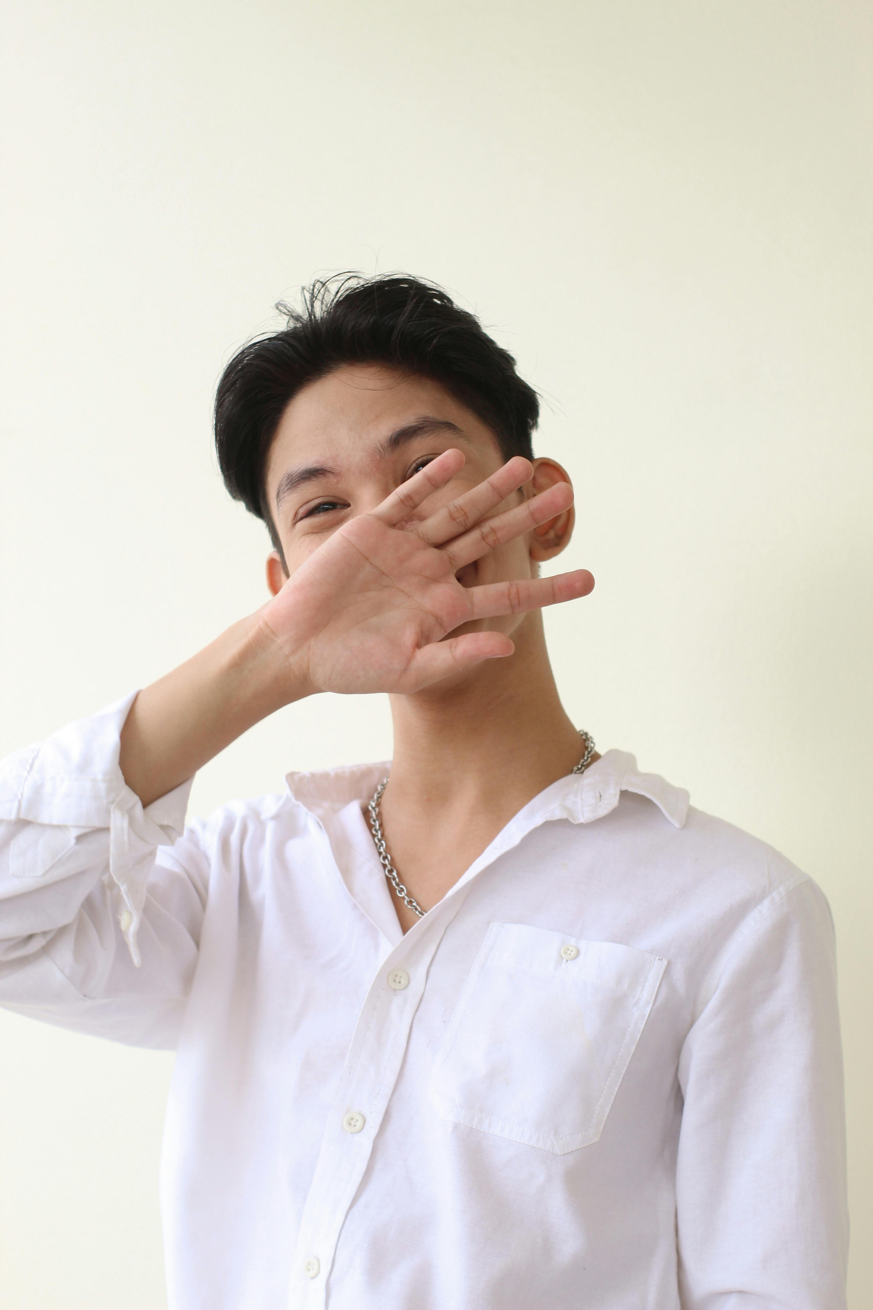 A young man wearing a white shirt smiles playfully while partially hiding his face with his hand.