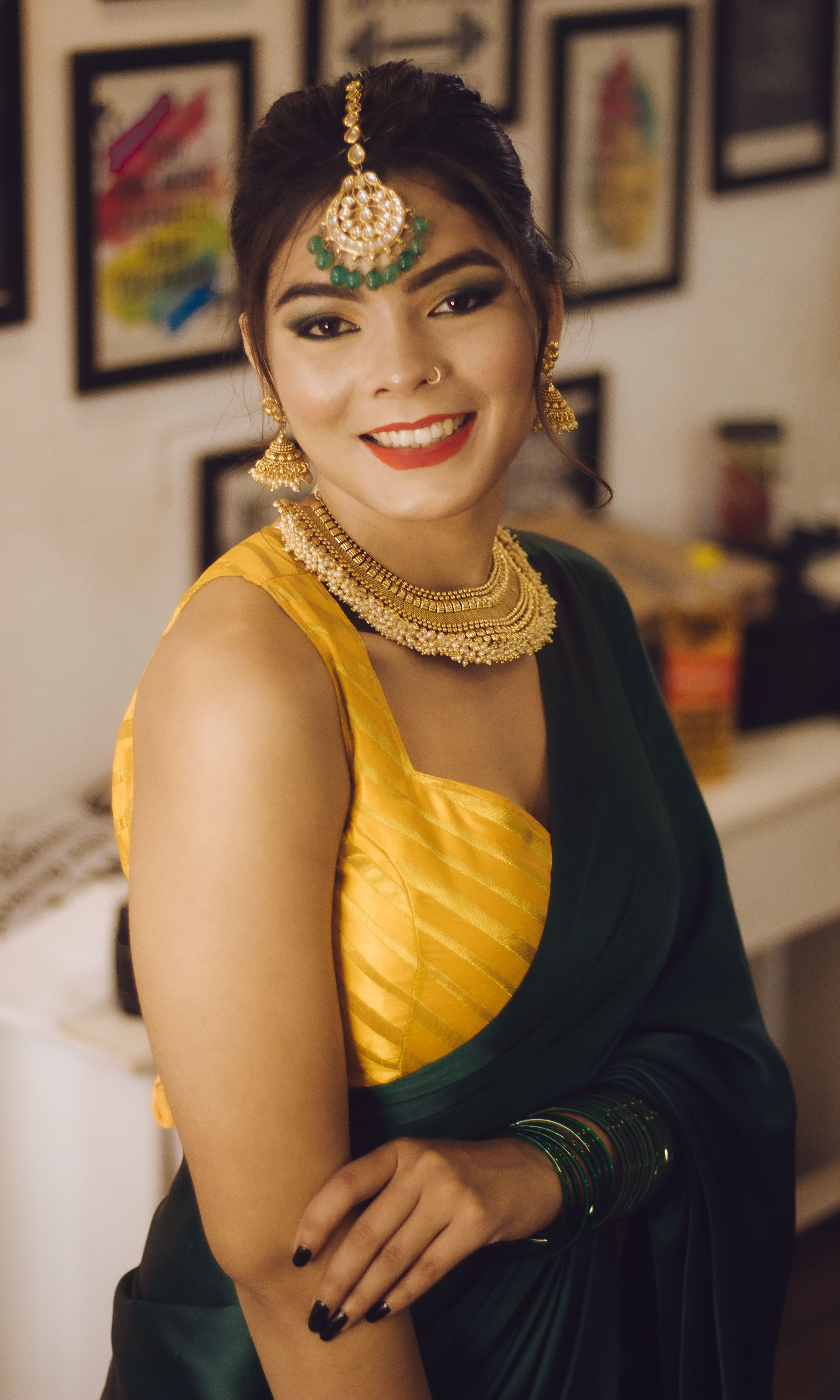 Smiling Indian woman in green and yellow sari with traditional jewelry, posing indoors.