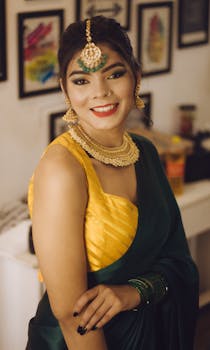Smiling Indian woman in green and yellow sari with traditional jewelry, posing indoors.