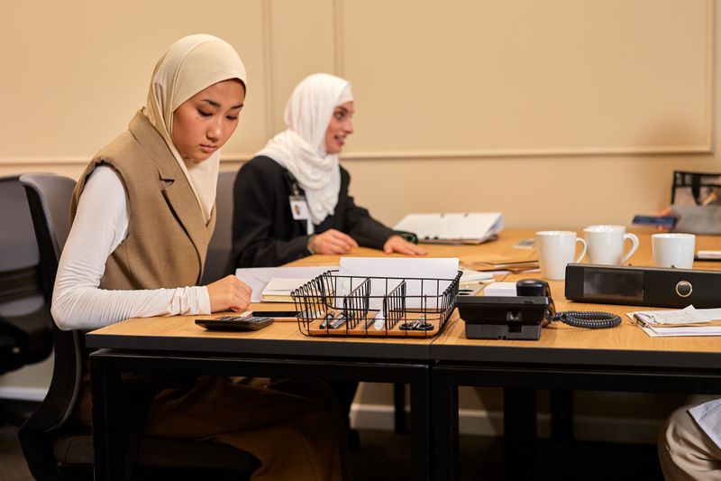 Two Muslim women in hijabs working diligently in a corporate office setting.