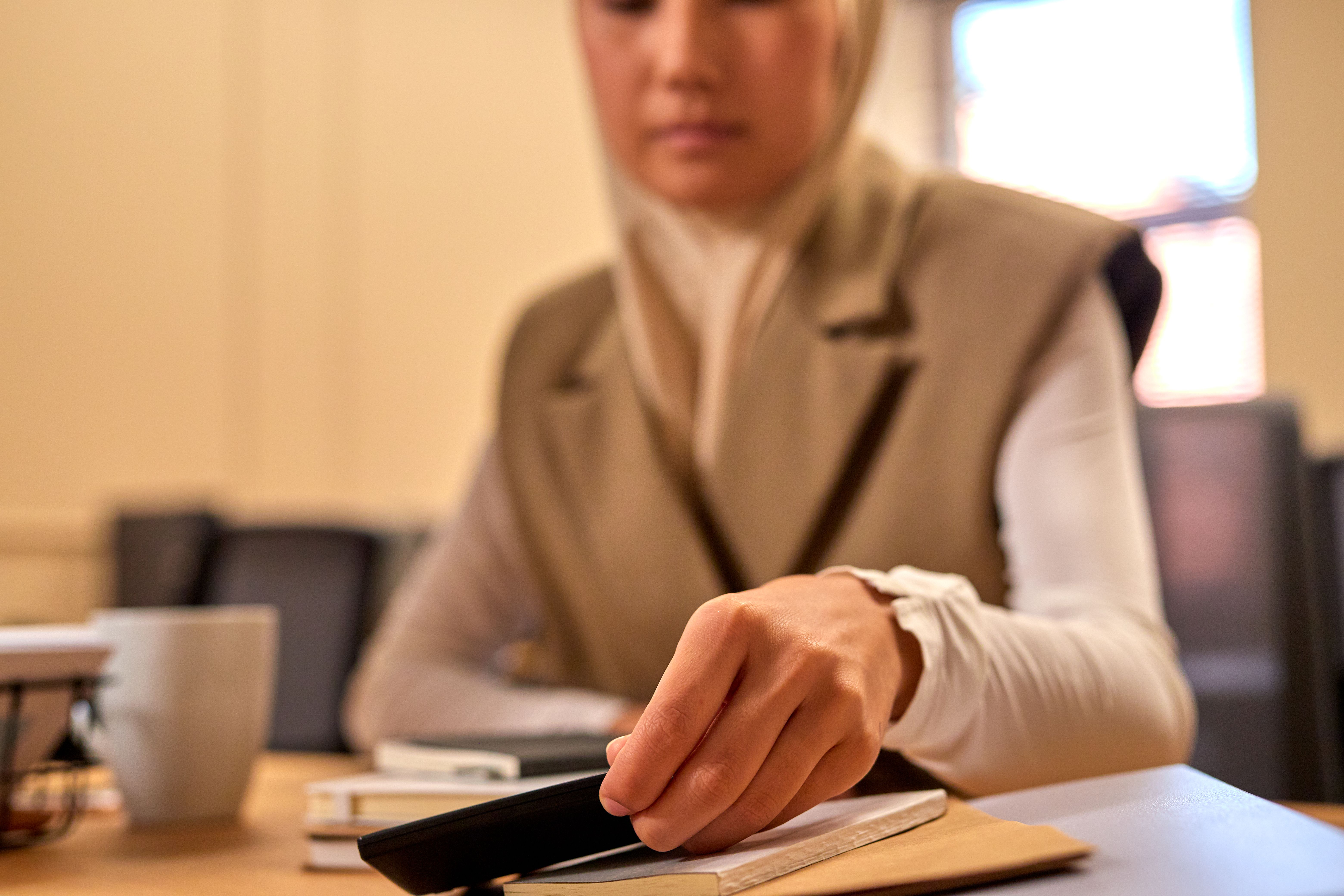 Mulher muçulmana em hijab utilizando smartphone enquanto está sentada em uma mesa interna, focando na tecnologia.