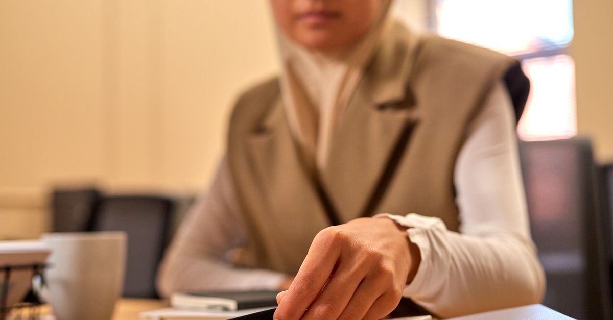 Mulher muçulmana em hijab utilizando smartphone enquanto está sentada em uma mesa interna, focando na tecnologia.