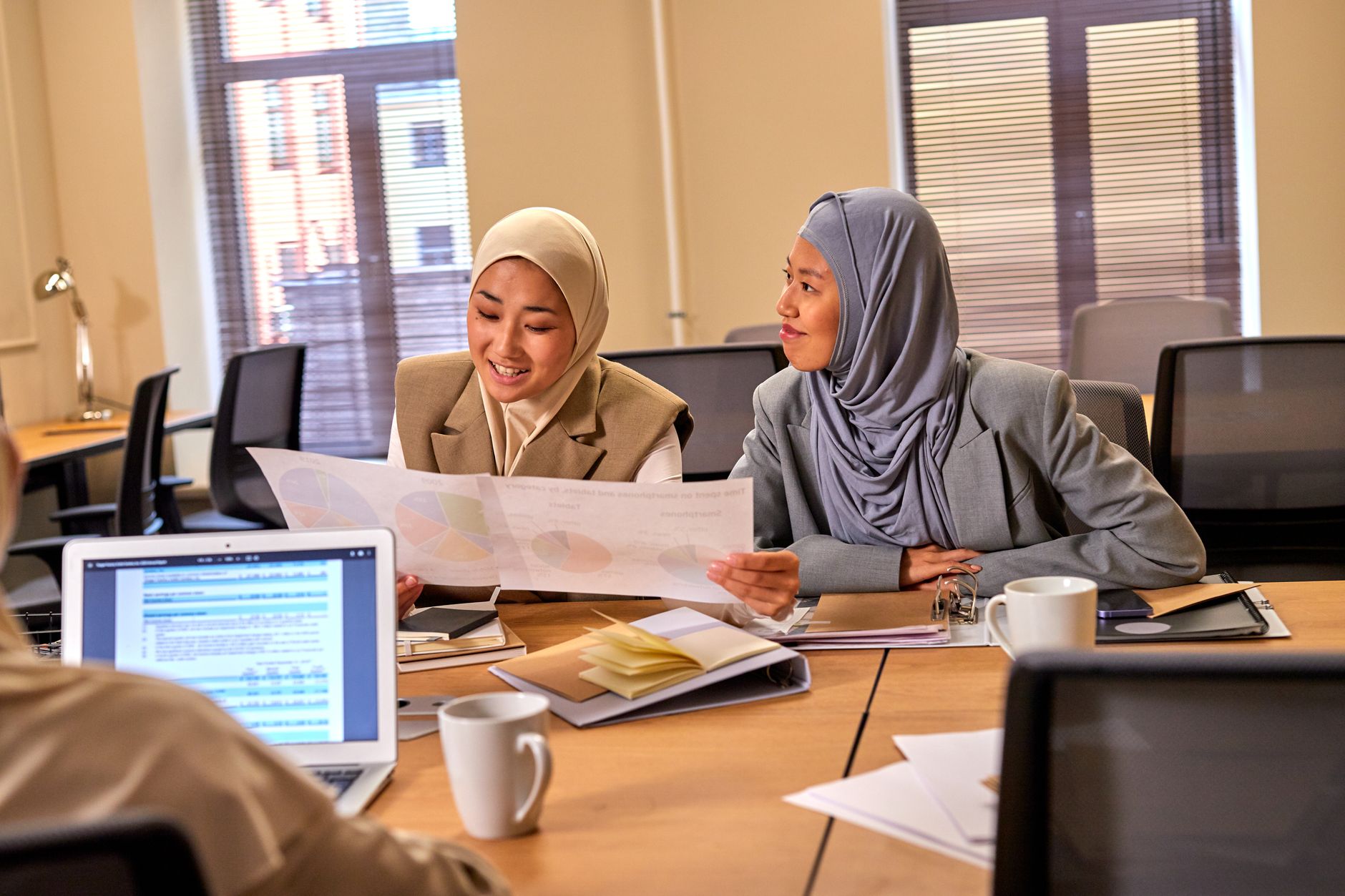 Two female colleagues in hijabs discussing charts in a sunny office setting, emphasizing diversity.