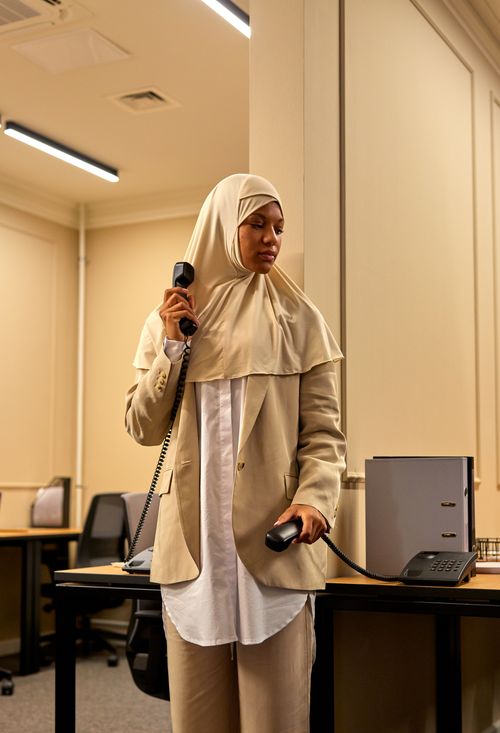 Free Businesswoman in beige hijab and suit using a landline phone in a modern office setting. Stock Photo