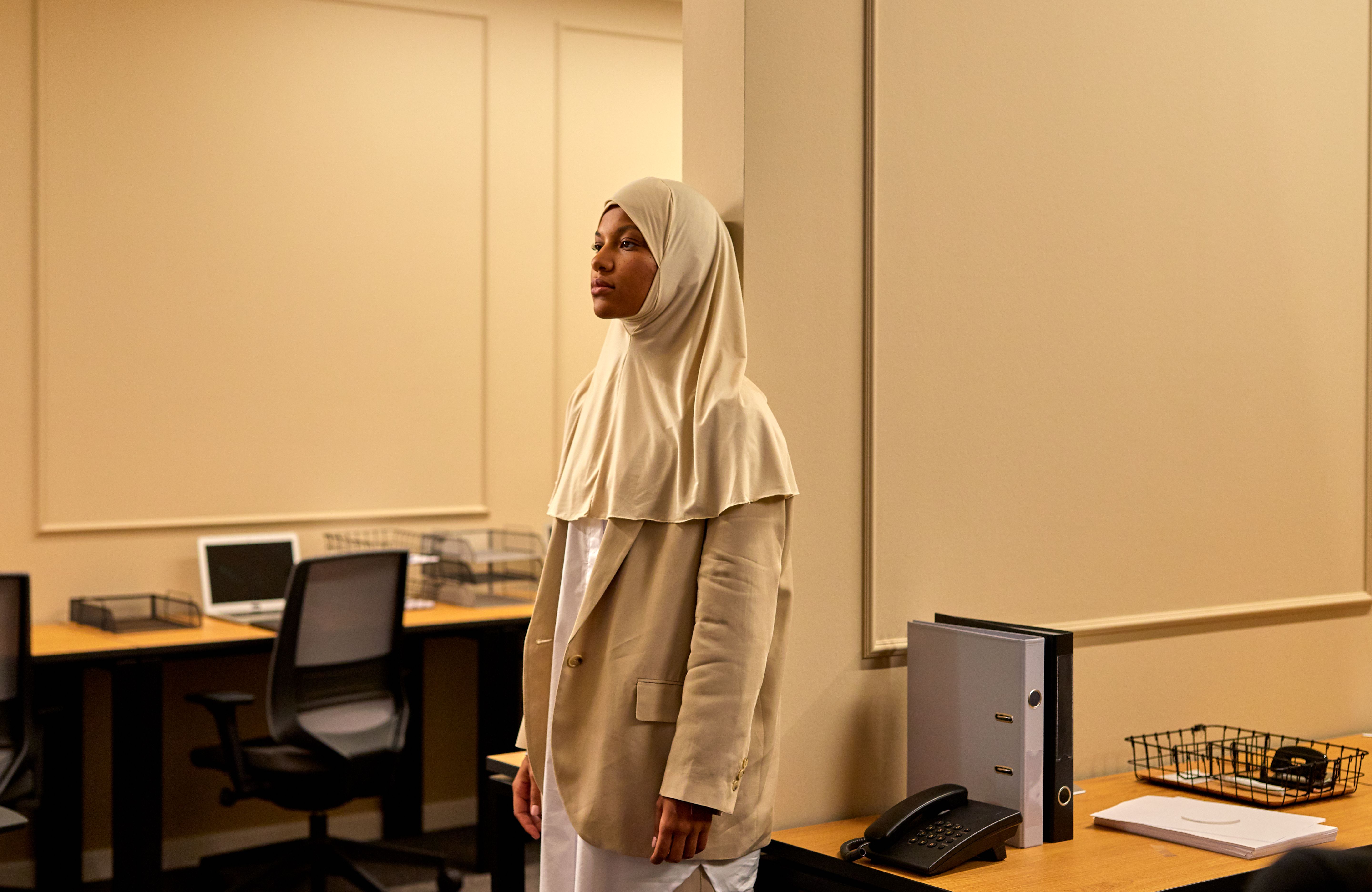 A woman in a hijab stands thoughtfully leaning against a wall in a modern office setting.
