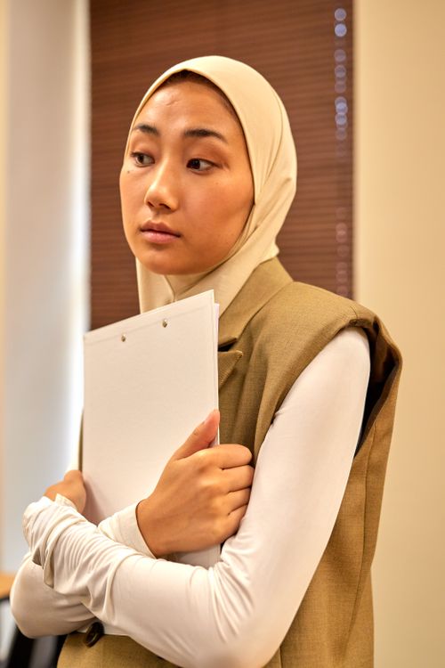 Free A young woman wearing a hijab holds a clipboard in a modern office setting. Stock Photo