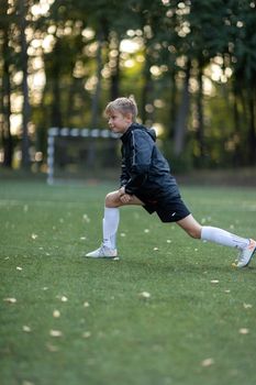 Boy in sportswear stretching on a soccer field amidst lush trees, preparing for a game.