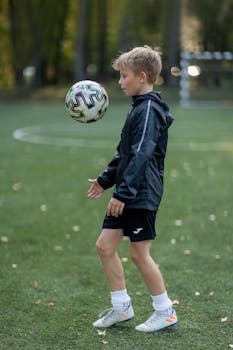 A young boy in sportswear practices soccer skills on a green football field during the day.
