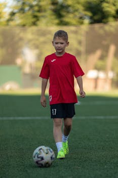 A young boy in a red jersey playing soccer on a green field during the day.
