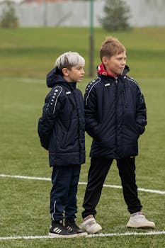 Two boys in jackets stand on a grass sports field, dressed warmly for cool weather.