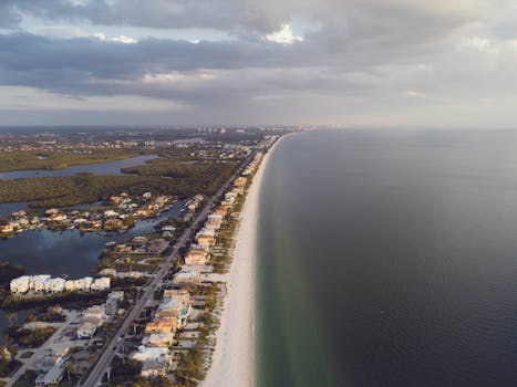 Stunning aerial view of coastal homes lining a serene beach with the ocean and clouds in the distance.