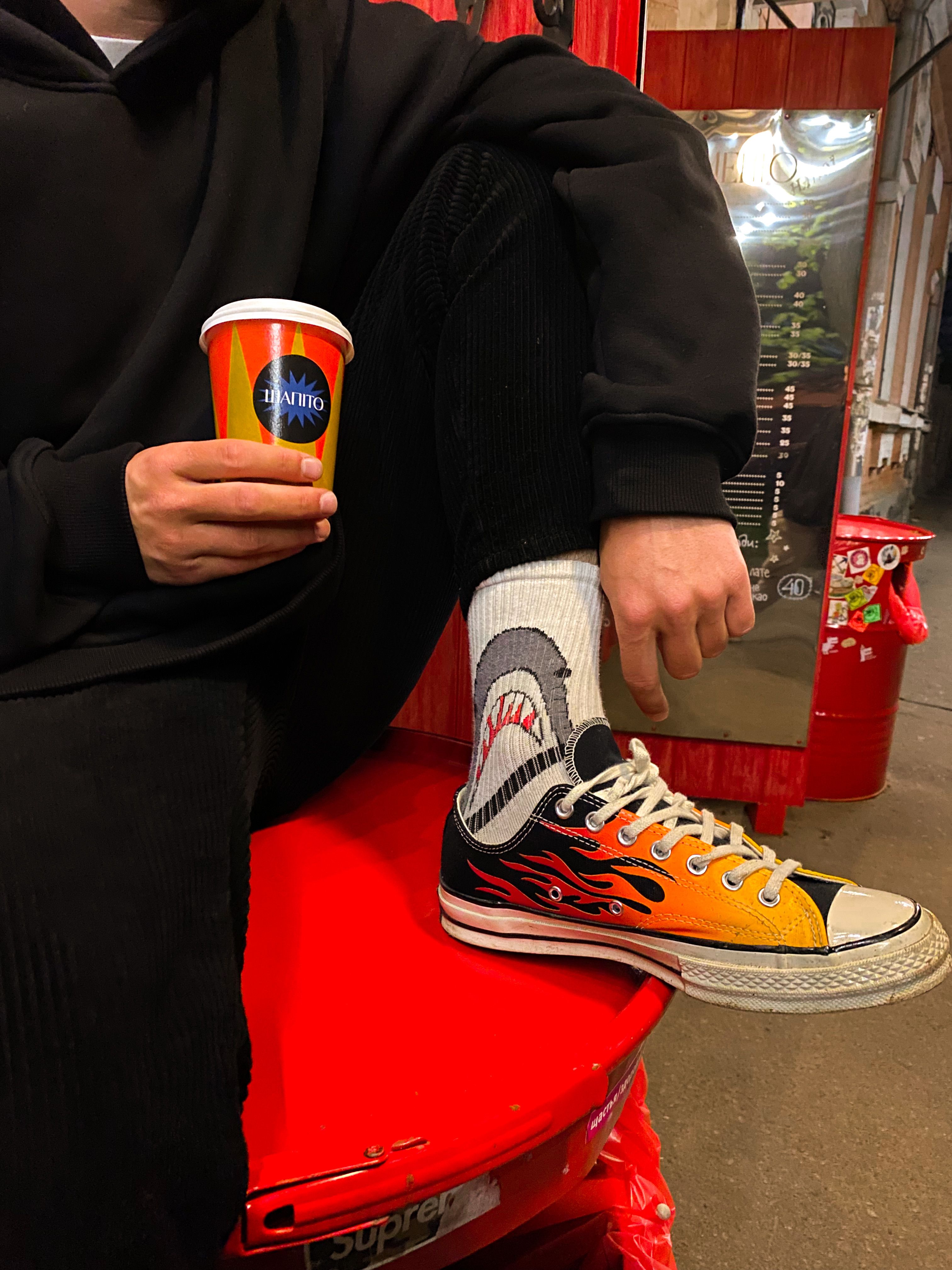 A person sitting with a colorful sneaker and shark sock holding a cup, showcasing urban style.